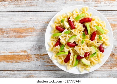 Delicious Mini Salami Bowtie Pasta Warm Salad With Avocado Slices And Basil Leaves On White Plate On Old Wooden Background,  View From Above