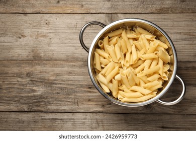 Delicious penne pasta in colander on wooden table, top view. Space for text