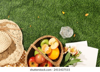 Delicious picnic spread with basket of fruits, hat, cup of cool water and white notebooks on green grass background. Top view, holiday summer concept. Space for design - Powered by Shutterstock