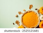 Delicious pumpkin pie on a plate, surrounded by autumn leaves, nuts, and mini pumpkins on a pastel green background. Thanksgiving and fall-themed culinary concept.