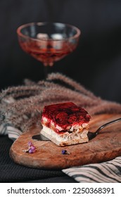 Delicious raspberry fruit cake next to a flower and glass of rose red dessert wine placed on light wooden board. Dark grey background. Served on a stainless steel cake spatula.