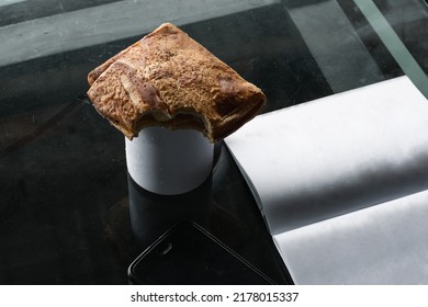 Delicious Sandwich Nibbled On Top Of A White Coffee Cup, Next To A Book With Its Blank Pages And On A Glass Table In The Company's Office. Light Breakfast In The Office Before Going To Work.