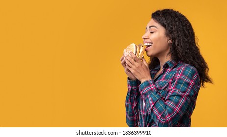 Delicious Sandwich. Side View Portrait Of Happy Black Female Model Eating Burger. African American Lady With Open Mouth Holding And Biting Snack, Orange Studio Background, Banner, Copy Space, Panorama