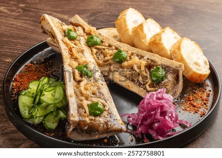 A delicious serving of bone marrow topped with herbs, accompanied by toasted bread, pickled onions, and sliced cucumbers, presented on a black plate against a wooden background.