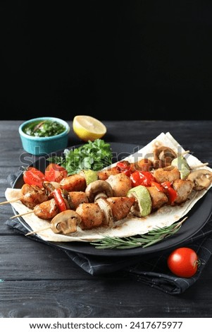 Delicious shish kebabs with vegetables served on black wooden table against dark background, space for text