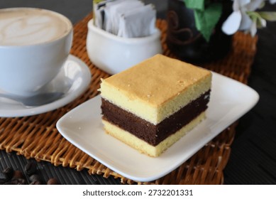 Delicious slice of layered cake featuring chocolate and vanilla, served on a white plate alongside a cup of latte, sugar packets, and roasted coffee beans, creating a delightful dessert setting