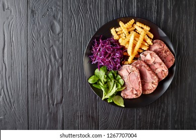 delicious sliced terrine of beef tongue and meat served with french fries, green leaves and red cabbage salad on a black plate on a wooden table, view from above, flat lay