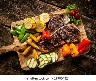 Delicious steakhouse porterhouse steak and colorful fresh roast vegetables with mangetout peas, corn, zucchini, bell pepper, potato and tomato on a wooden board, view from above