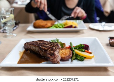 Delicious Strip Loin Beef Steak Meal Served With Fresh Vegetables And Marsala Portobello Mushroom Sauce. Focus On Steak And Deliberate Shallow Depth Of Field On Subject Having A Meal Across The Table.