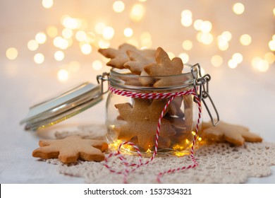 Delicious Sweet Christmas Gingerbread Cookies In Glass Jar Decorated With Red Ribbon. Best Homemade Present For Family! Lights On Background, White Table.