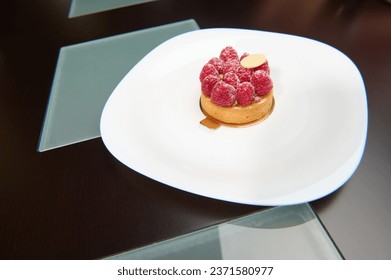 A delicious sweet tartlet with fresh ripe organic raspberries on a white plate over wooden background. French patisserie and boulangerie. Food and drink consumerism. Copy advertising space
