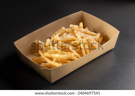 Delicious, tasty french fries topping with Parmesan cheese on top on a black background, in a container for delivery. Deep fry potato or appetizer, fast food. Dish of crisp golden potato chips.
