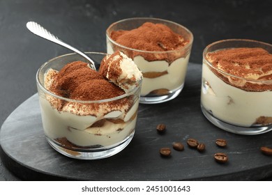 Delicious tiramisu in glasses, spoon and coffee beans on black table