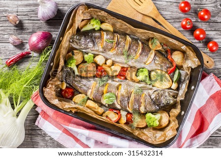 delicious trout fish baked with potatoes, broccoli, lemon, tomatoes and spices in baking dish on a wooden background, top view