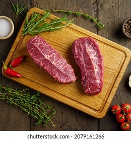 Delicious Two Raw Top Blade Beef Steak On Wooden Chopping Board With Spices And Herbs. Cherry Tomatoes, Thyme, Rosemary, Salt, Pepper, Garlic Clove, Red Chili. Overhead, Closeup