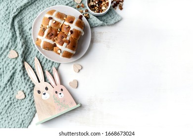 Delicious vegan homemade hot cross buns on a plate. The plate sits on a sage coloured tea towel and is surrounded by a bowl of various nuts, wooden hearts and wooden easter bunny decorations. 