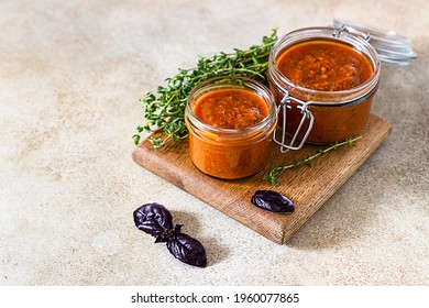 Delicious vegetable caviar in jars, thyme and purple basil on wooden board, light stone background. Homemade caviar with eggplant, tomatoes and bell pepper.