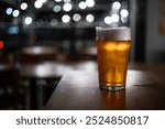 A delicious-looking pint of top-fermented beer sits on a rustic wooden table in a pub. The background features bokeh circles from the lights, creating a warm and inviting atmosphere.