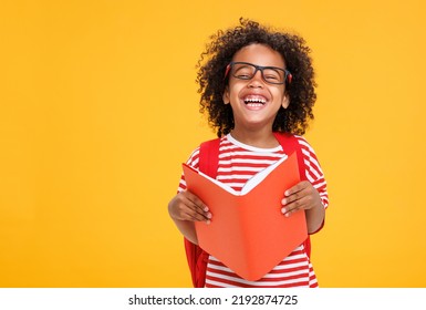 Delighted African American boy in glasses laughing for camera and reading book  while having fun during school students against yellow background - Shutterstock ID 2192874725