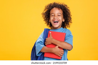 Delighted African American boy in glasses laughing for camera and reading book  while having fun during school students against yellow background