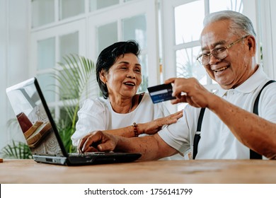 Delighted Asian man and woman using laptop and credit card to make order in online shop while sitting at table at home. Senior couple doing online shopping