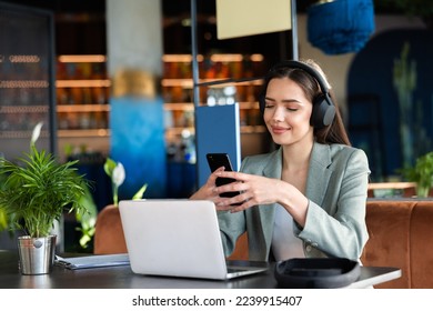 Delighted attractive young buisness woman working from caffe online using digital devices laptop cellphone and earphones to comunicate and havung conversation. Working process concept.