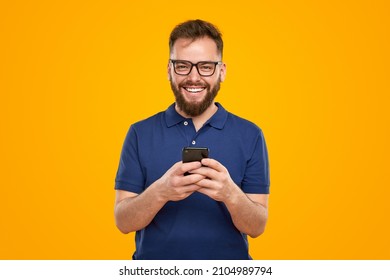 Delighted bearded male in glasses and blue t shirt messaging on social media, via mobile phone while looking at camera on yellow background in studio