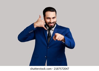 Delighted Bearded Man Standing With Telephone Hand Gesture And Smiling To Camera, Flirting Offering To Contact By Phone, Pointing Finger To Camera. Indoor Studio Shot Isolated On Gray Background.