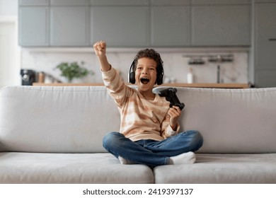 Delighted black boy in headphones holding game controller and raising fist in victory, happy african american male child enjoying playing video game, sitting on sofa in bright living room at home - Powered by Shutterstock