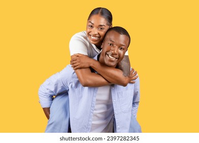 Delighted black couple enjoying piggyback ride, woman laughing and embracing the man from behind, set against bright yellow background