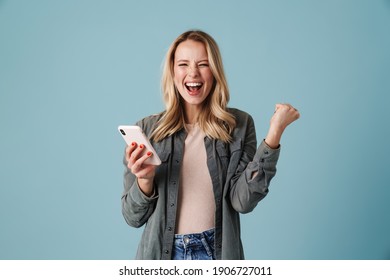 Delighted Blonde Girl Making Winner Gesture And Using Mobile Phone Isolated Over Blue Background