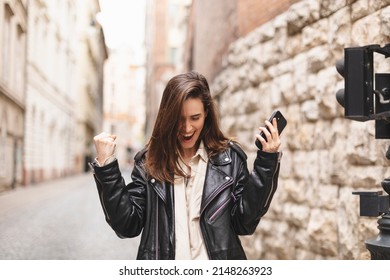 Delighted brunette girl making winner gesture and using mobile phone walking on the street. Portrait of surprised hipster girl surfing internet on mobile phone outdoors. 