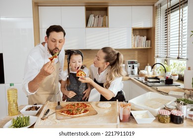 Delighted curious child kid daughter biting piece of homemade pizza. Mother feeding daighter with hands. Happy father with beared tasting pizza. Pizza ingredients on wooden table for baking cooking. - Powered by Shutterstock