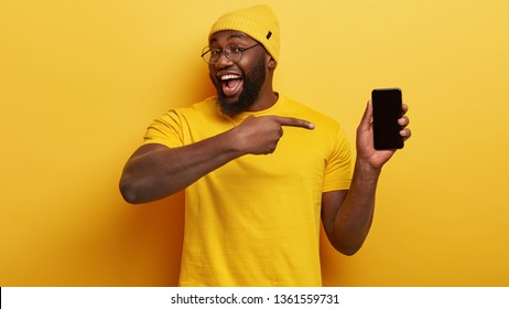 Delighted dark skinned male seller of modern devices, points at mobile phone with mock up black screen suggests buying gadget for low price wears yellow fashionable clothes in one tone with background