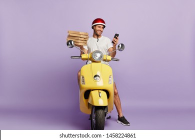 Delighted Delivery Man Checks Route Via Special Application On Mobile Phone, Holds Paper Containers With Pizza, Delivers Fast Food, Wears Protective Helmet, Rides Yellow Motorbike, Isolated On Purple