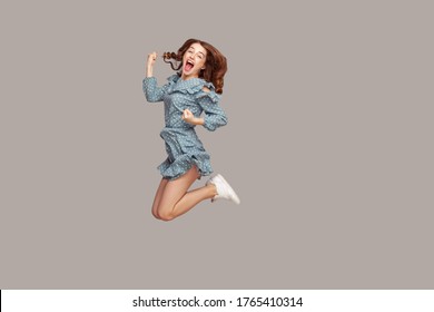 Delighted enthusiastic girl ruffle dress flying mid-air with raised fists shouting for joy, jumping trampoline looking at camera extremely happy, celebrating success. studio shot isolated on gray