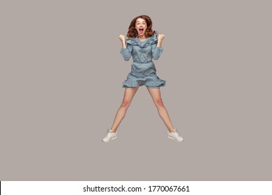 Delighted enthusiastic girl ruffle dress flying mid-air with raised fists shouting for joy, jumping trampoline looking at camera extremely happy, celebrating success. studio shot isolated on gray