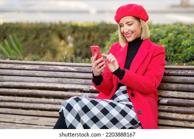 Delighted Female In Red Coat And Beret Scrolling News On Modern Cellphone While Sitting On Wooden Bench In Autumn Park