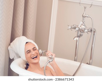 Delighted female with towel on head lying in soaker bathtub and imitating conversation on phone call with shower water can - Powered by Shutterstock