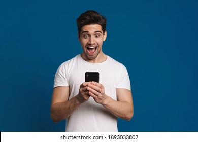 Delighted guy expressing surprise while using mobile phone isolated over blue background