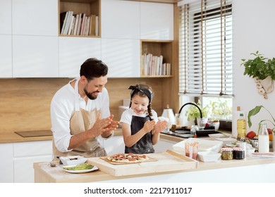 Delighted kid with father in aprons clapping in ahnds looking at selfmade pizza on wooden table. Preparing to put pizza in oven. Delighted parent having fun with beautiful daughter with braids.