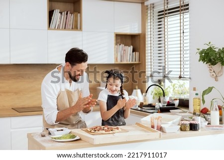 Delighted kid with father in aprons clapping in ahnds looking at selfmade pizza on wooden table. Preparing to put pizza in oven. Delighted parent having fun with beautiful daughter with braids.