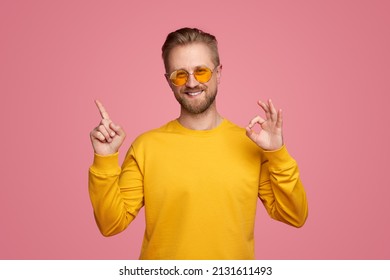 Delighted male hipster showing okay gesture and pointing up with index finger while smiling and looking at camera against pink background in studio