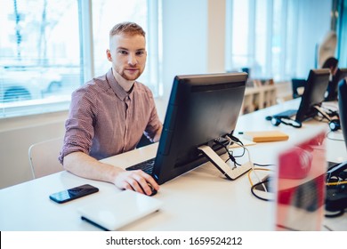 Delighted Male In Stylish Shirt Sitting At Table In Office And Using Modern PC Computer While Looking At Camera On Blurred Background. Online Courses At School