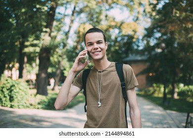 Delighted male with toothy smile and earphones looking at camera while standing in park and having conversation on mobile phone