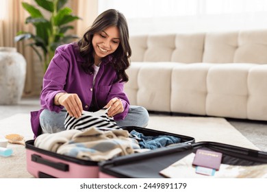 Delighted middle eastern woman kneeling and packing a variety of clothes into her travel luggage, smiling warmly