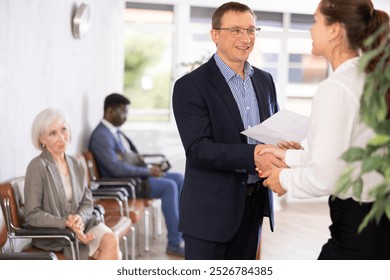 Delighted middle-aged man with a document in one hand shaking hands with a person against the background of visitors sitting in waiting room
