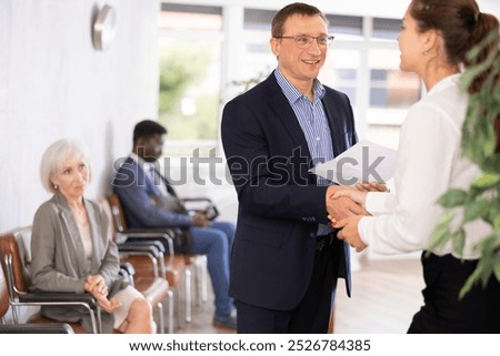 Delighted middle-aged man with a document in one hand shaking hands with a person against the background of visitors sitting in waiting room