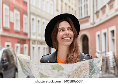 Delighted optimistic female toursit finds place on map, walks in city center during summer trip, wears stylish black hat, poses against urban setting, checks out sights, tours abroad, plans route
