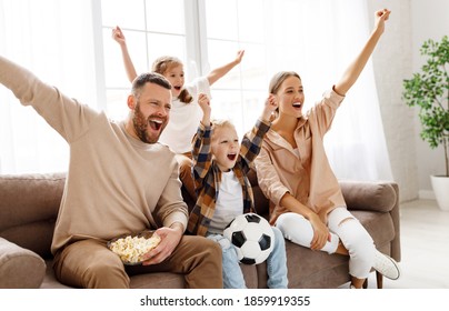 Delighted parents and children with popcorn and ball raising arms and screaming while watching football match and celebrating goal at home